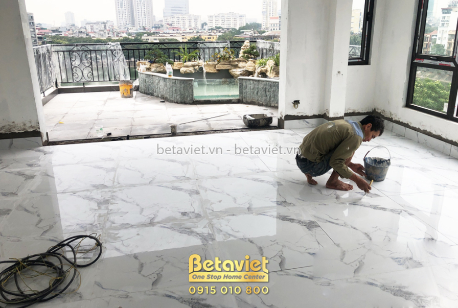 Công nhân đang lát gạch sàn màu trắng vân đá tại một không gian tầng trên, có cửa kính lớn nhìn ra ban công và cảnh quan t…