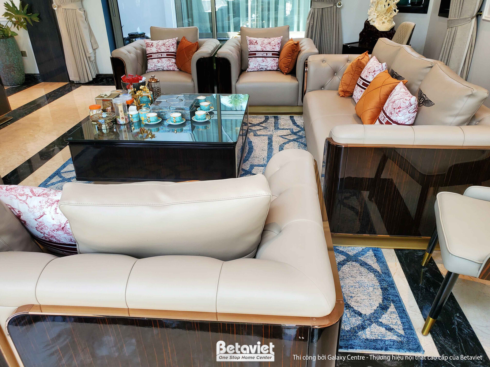 Cận cảnh bộ sofa da màu kem với đường nét bo cong mềm mại, gối tựa màu cam đất tạo điểm nhấn. Bàn trà mặt kính đen với khu…