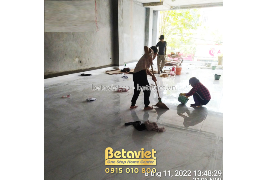 Không gian tầng trệt đang được vệ sinh sau khi thi công phần thô. Sàn bê tông còn bụi bẩn, công nhân đang quét dọn. Ánh sá…