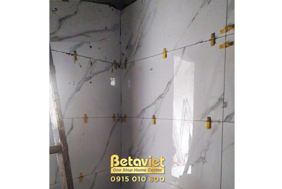 Cận cảnh công nhân đang ốp lát gạch tường màu trắng vân mây. Các miếng gạch được cố định bằng ke nhựa, đảm bảo độ phẳng và…