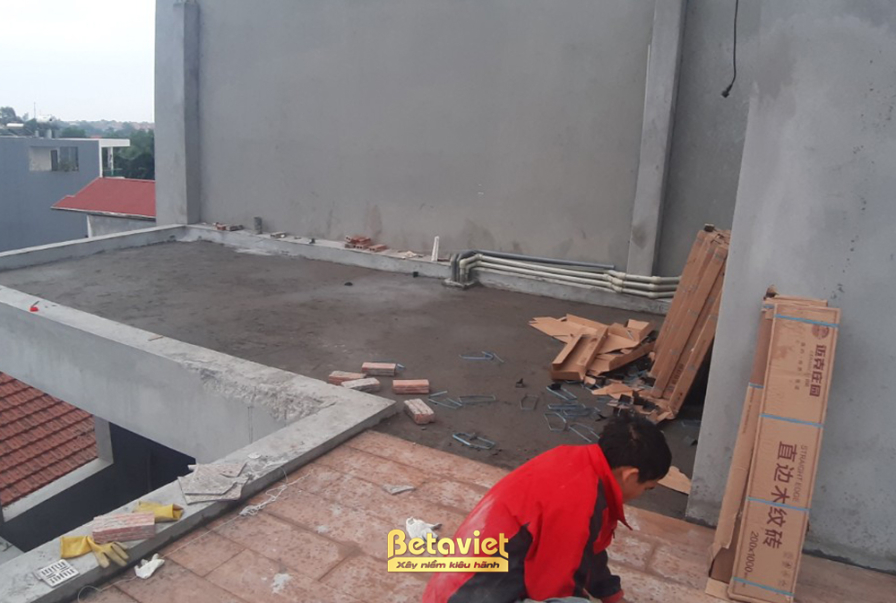Tầng thượng với sàn bê tông xám đã hoàn thiện, mái ngói đỏ của nhà liền kề nhìn thấy phía xa. Vật liệu sàn gỗ còn đóng tro…