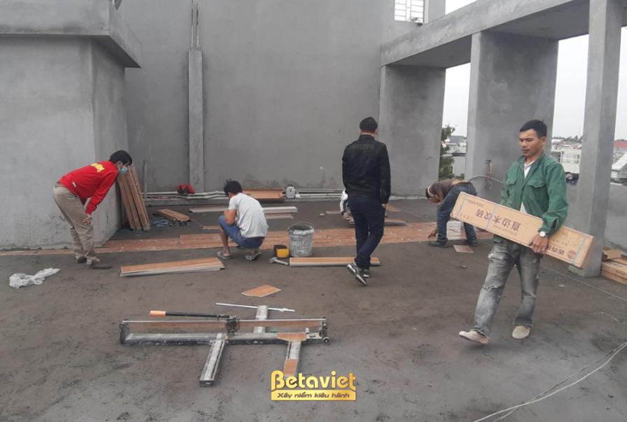 Không gian nội thất đang trong giai đoạn lát sàn gỗ, với các công nhân đang vận chuyển và lắp đặt từng tấm ván gỗ. Bố cục…