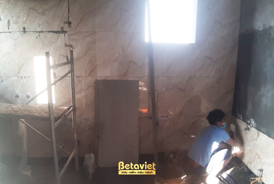 Không gian nhà vệ sinh đang trong giai đoạn ốp lát sàn, với một công nhân đang tỉ mỉ đặt từng viên gạch. Bố cục ảnh tập tr…