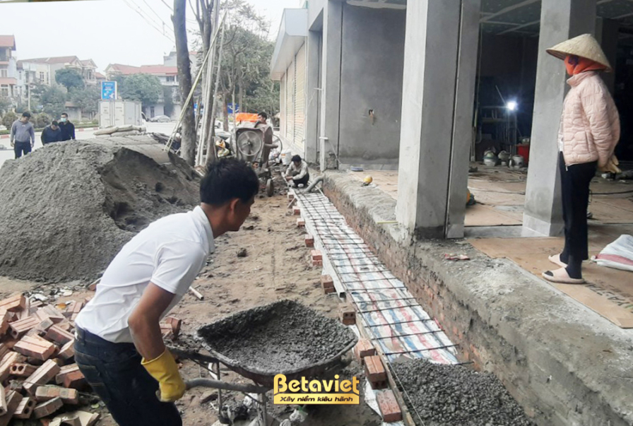 Góc nhìn từ bên ngoài vào mặt tiền công trình nhà phố đang xây dựng, với các bức tường gạch đỏ và khung cửa sổ lớn. Giàn g…