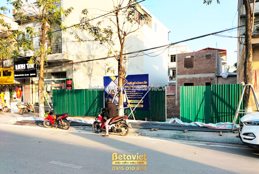 Toàn cảnh công trường đang thi công phần móng và tầng trệt từ góc nhìn khác. Giàn giáo và lưới bảo hộ bao quanh. Cây xanh…