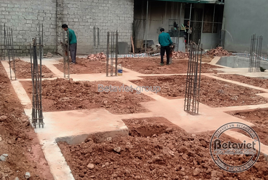 Khu đất móng nhà phố đang đào đất, nền đỏ lộ rõ, hệ cột thép chờ dựng thẳng hàng chuẩn bị đổ móng. Công nhân đo đạc định v…