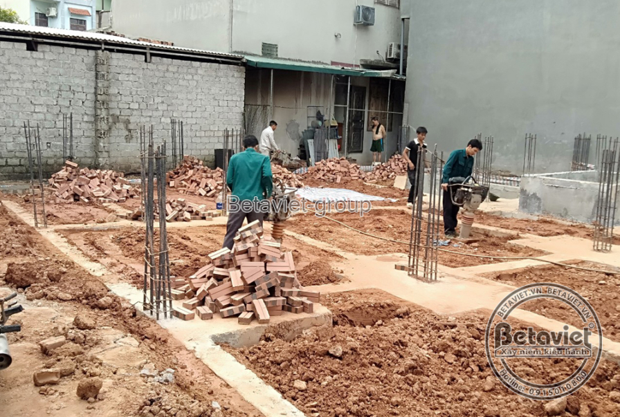 Công tác thi công móng nhà đang diễn ra, với các công nhân đang làm việc trên hệ thống cốt thép và cốp pha gạch. Bố cục ản…