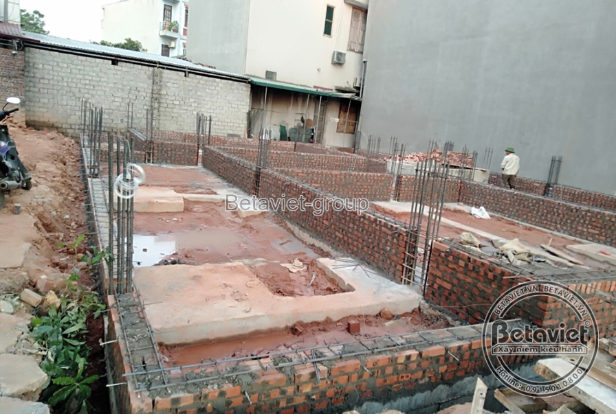 Cận cảnh công tác thi công móng nhà, với hệ thống cốt thép và cốp pha gạch được đặt trong nền đất. Bố cục ảnh tập trung và…