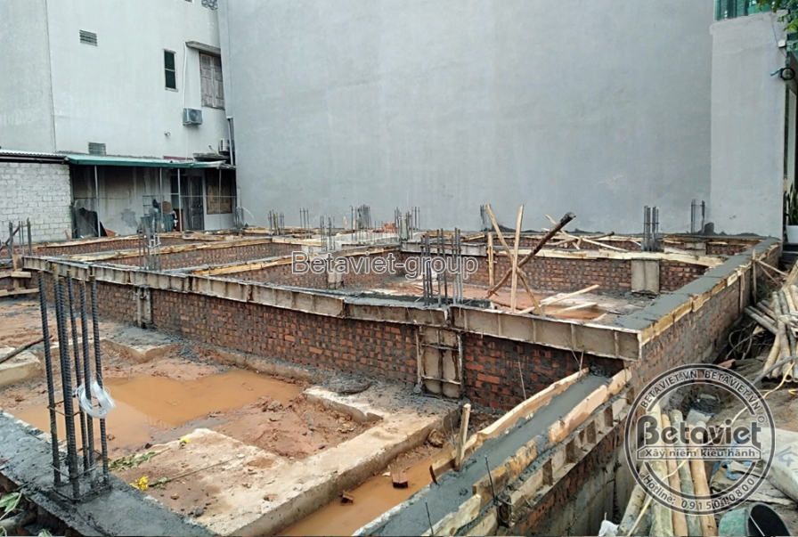 Cận cảnh công tác thi công móng nhà, với hệ thống cốt thép và cốp pha gạch được đặt trong nền đất ẩm. Bố cục ảnh tập trung…