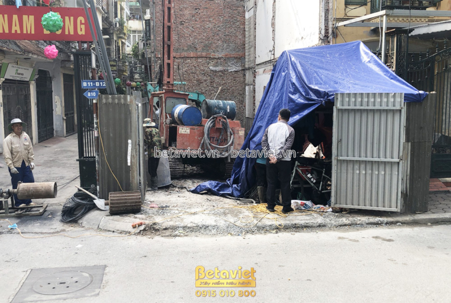 Công trường nhà phố nhìn từ đường phố, với các công nhân đang làm việc và vật liệu xây dựng chất đống. Có bạt che và biển…