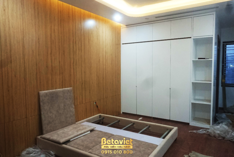 Phòng ngủ đang lắp đặt giường bục thấp bằng gỗ màu nâu trầm, kết hợp với vách ốp gỗ màu vàng nhạt. Tủ quần áo âm tường cán…