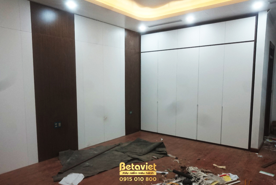 Phòng ngủ với hệ tủ quần áo âm tường lớn cánh phẳng màu trắng, chiếm trọn một mảng tường. Vách tường đối diện ốp gỗ màu nâ…