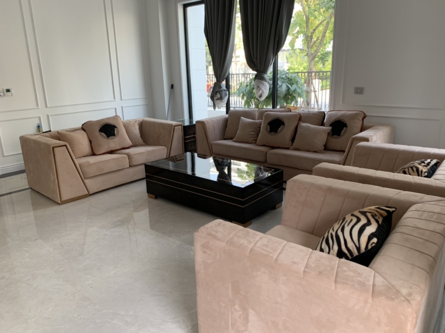 Phòng khách rộng rãi với bộ sofa bọc nỉ màu kem trung tính, gồm hai sofa dài và hai ghế đơn, bố cục đối xứng. Bàn trà trun…