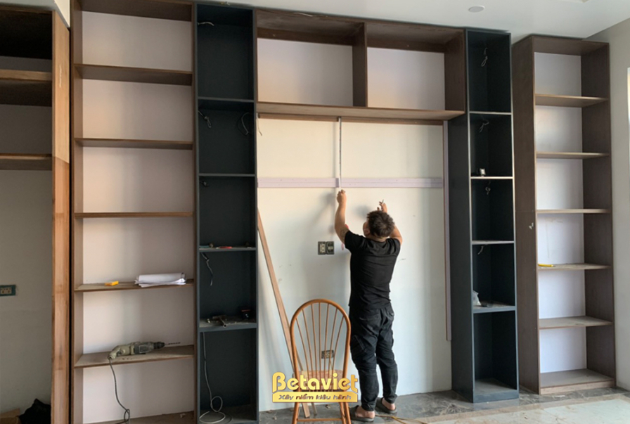 Công nhân đang lắp đặt các đợt kệ cho hệ tủ sách hoặc tủ trưng bày lớn. Các ngăn kệ được thiết kế đa dạng, kết hợp giữa gỗ…