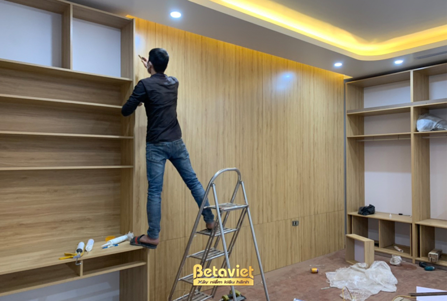 Công nhân đang đứng trên thang để lắp đặt các tấm ốp và kệ gỗ cho hệ tủ lớn ốp tường. Vách tường ốp gỗ màu vàng nhạt, kết…