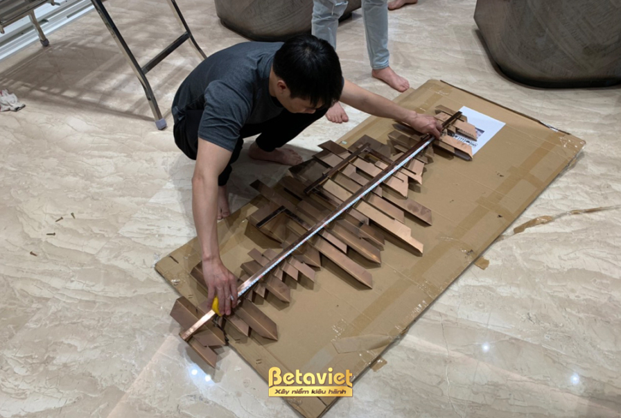 Cận cảnh đội ngũ kỹ thuật Betaviet đang đo đạc và căn chỉnh tỉ mỉ một chi tiết trang trí ốp tường bằng gỗ, thể hiện sự chu…