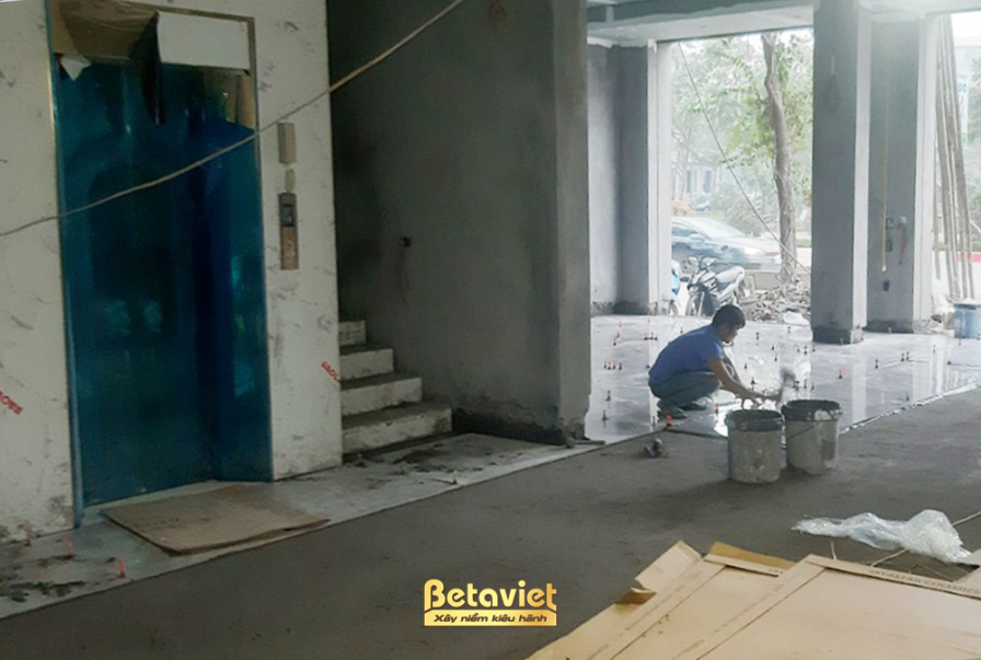 Không gian nội thất đang trong giai đoạn ốp lát sàn, với một công nhân đang trải vữa xi măng. Bố cục ảnh tập trung vào sàn…