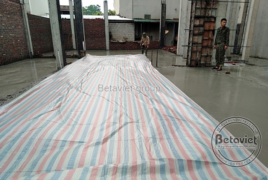 Không gian sàn bê tông cốt thép đã được đổ và phủ bạt, thể hiện tiến độ thi công phần thô. Bố cục ảnh từ trên cao, cho thấ…