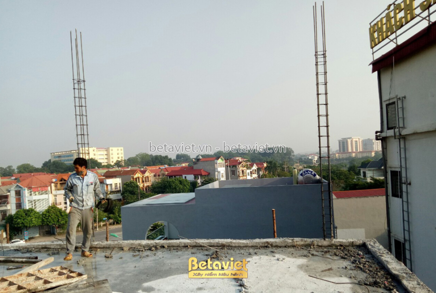 Không gian tầng mái đang được thi công, với các cột bê tông và thang kim loại. Bố cục ảnh từ trên cao, cho thấy sự phát tr…