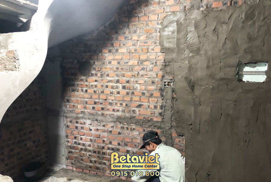Công nhân đang trát vữa lên bức tường gạch đỏ bên trong nhà. Bố cục cận cảnh, cho thấy rõ vật liệu gạch và lớp vữa mới đượ…