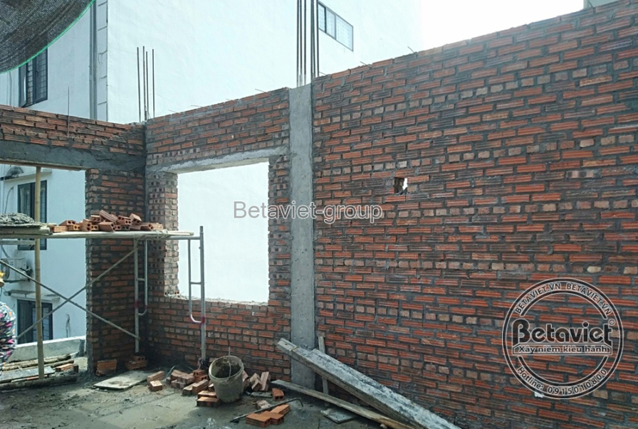 Mặt tiền công trình nhà phố đang trong giai đoạn xây thô, với các bức tường gạch đỏ và khung cửa sổ đã định hình. Bố cục ả…