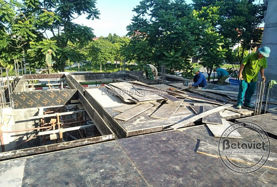 Công nhân đang làm việc trên sàn bê tông cốt thép đã được đổ, với cốp pha và giàn giáo xung quanh. Bố cục ảnh từ trên cao…