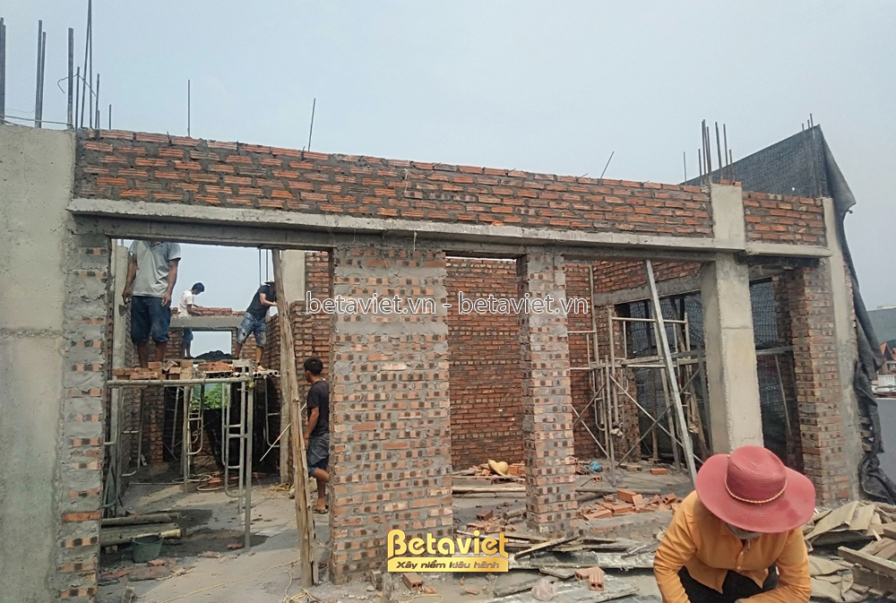 Tường gạch đỏ tầng 4 đang được xây dựng, giàn giáo gỗ bao quanh khung cửa sổ còn để trống. Công nhân đội mũ bảo hộ thao tá…