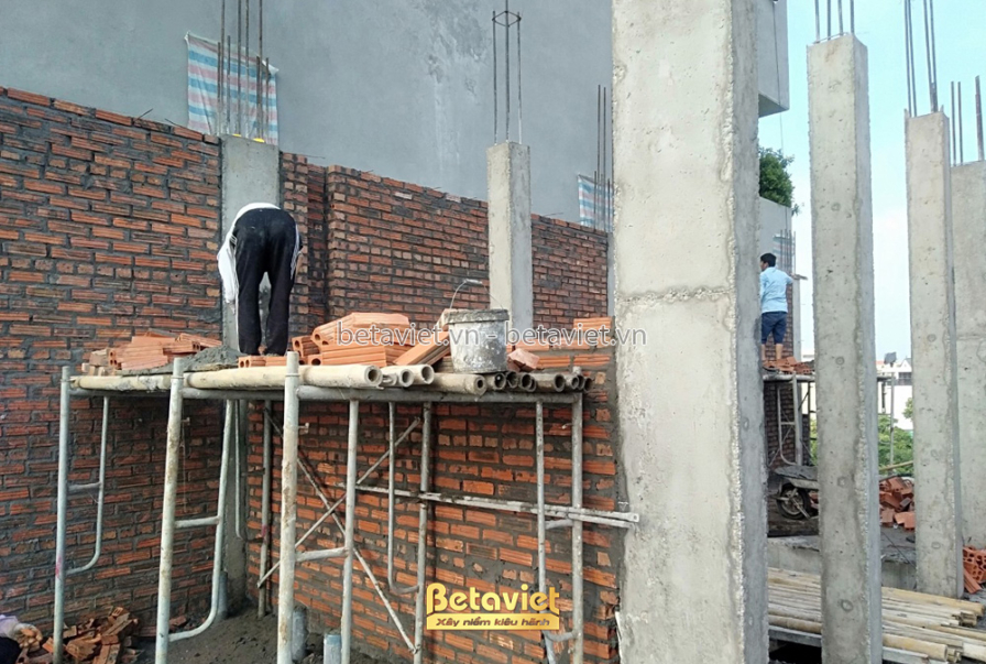 Công nhân đang xây tường gạch trên tầng 4 của công trình nhà phố, với giàn giáo và vật liệu xây dựng xung quanh. Bố cục ản…