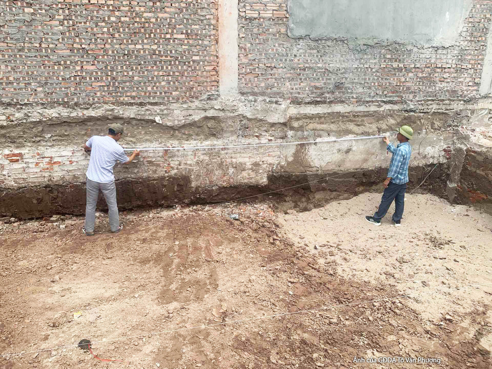 Hai công nhân đang kiểm tra khu vực móng hoặc tường bao của công trình, với lớp đất nền và gạch xây thô lộ rõ. Bối cảnh ch…