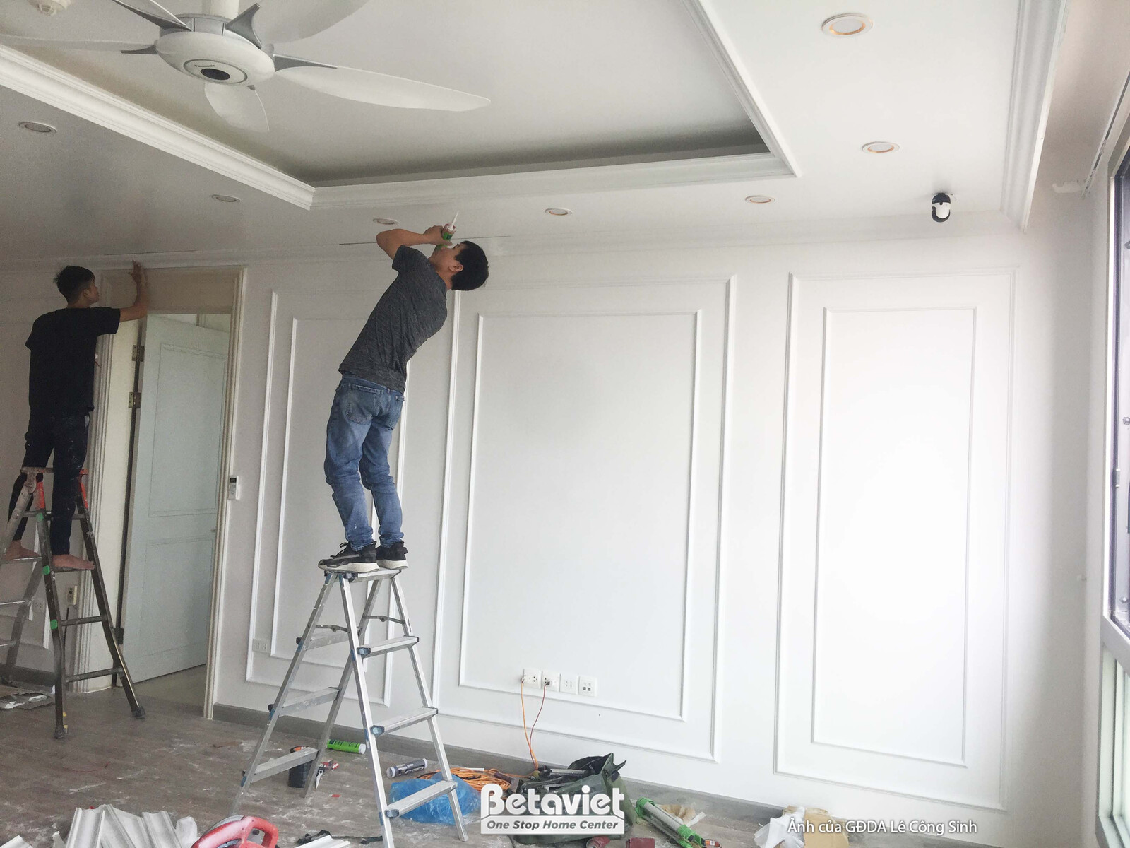 Cảnh thi công hoàn thiện chi tiết phào chỉ và lắp đặt hệ thống điện trên tường. Hai công nhân đang làm việc trên thang, tậ…