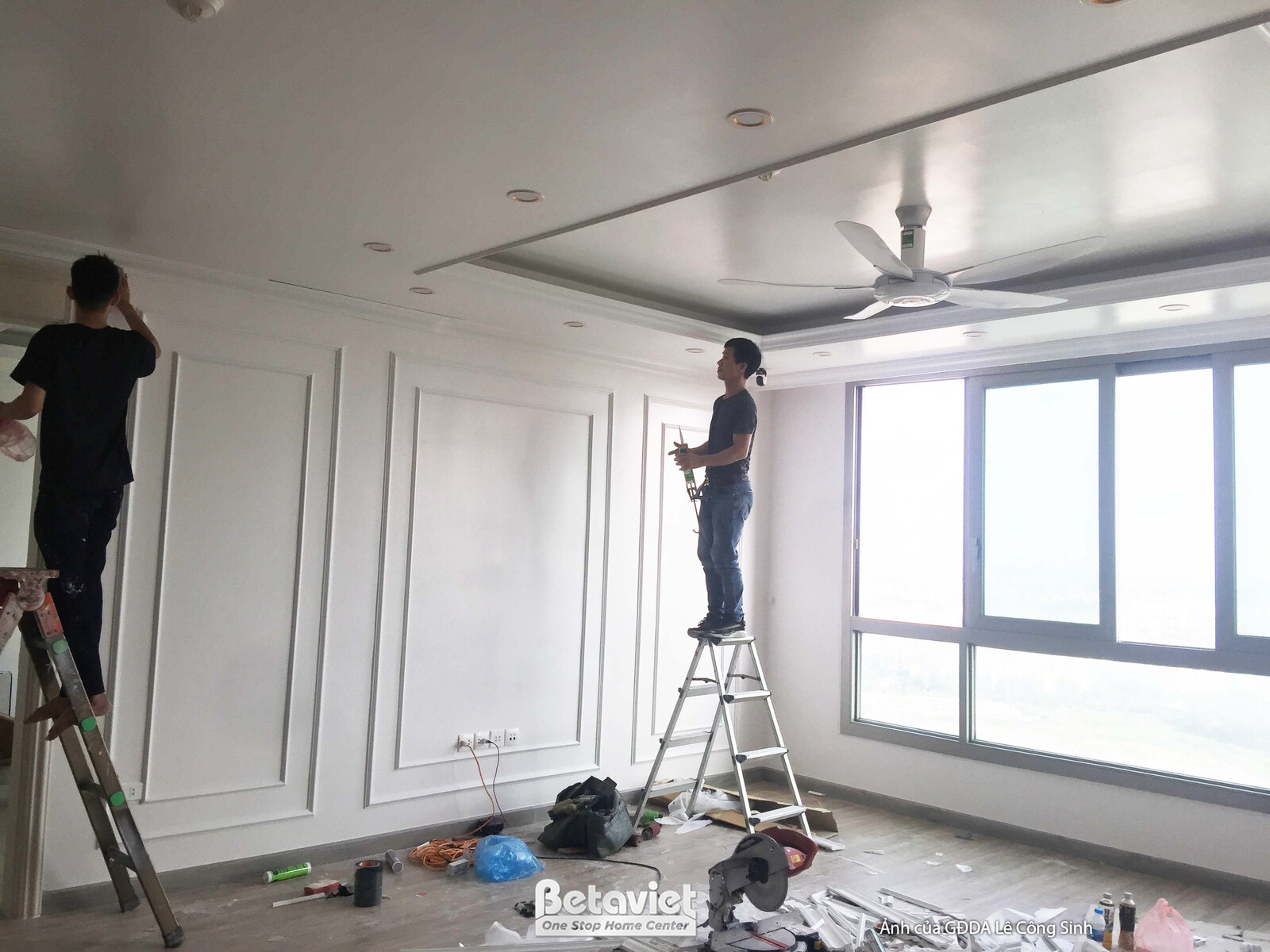 Một công nhân đang thực hiện công đoạn hoàn thiện phào chỉ trên tường trong không gian phòng khách. Tường được sơn màu trắ…