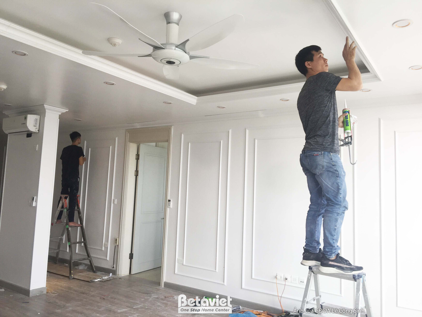 Một công nhân đang lắp đặt hoặc kiểm tra hệ thống đèn và quạt trần trong không gian phòng khách. Trần thạch cao màu trắng…