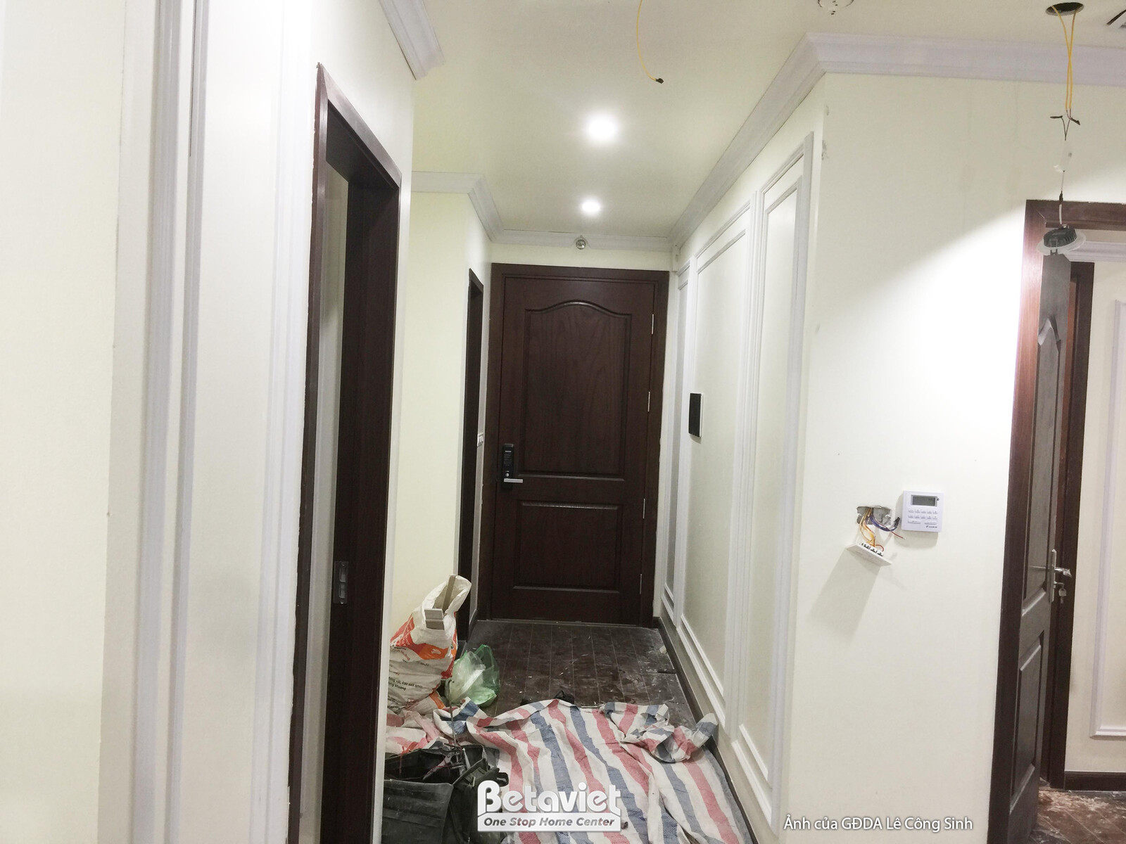 Lối vào căn hộ hoặc hành lang đang trong quá trình thi công, với tường được ốp phào chỉ màu trắng trang nhã. Cửa gỗ màu nâ…