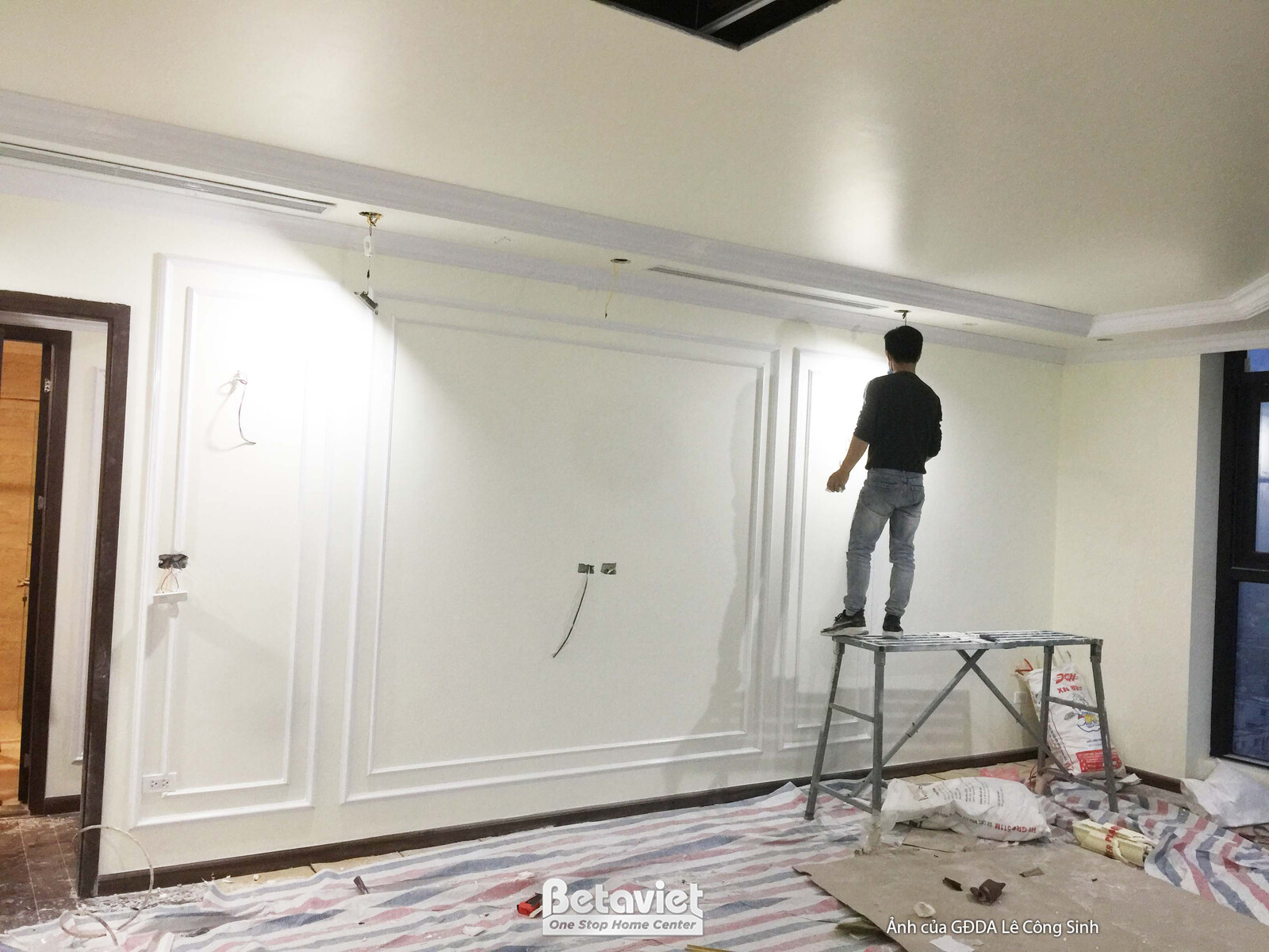 Người thợ đang tỉ mỉ hoàn thiện phần tường ốp phào chỉ màu trắng trong không gian phòng khách hoặc phòng sinh hoạt chung…