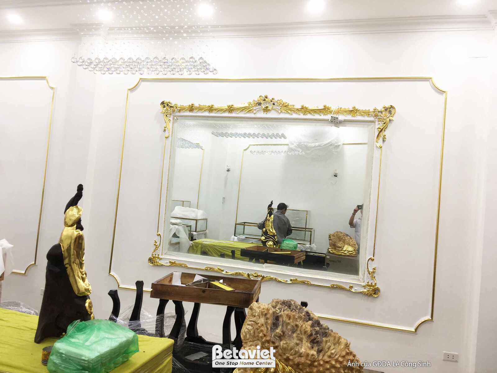 Chi tiết không gian sảnh với gương lớn viền vàng chạm khắc tinh xảo, phản chiếu một phần không gian cửa hàng. Tường trắng…