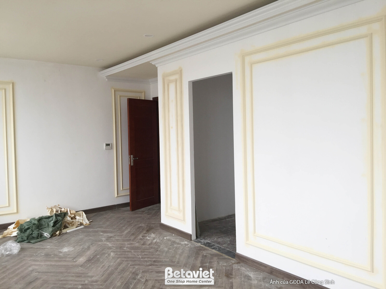 Khu vực hành lang hoặc lối đi đang thi công, với tường được trang trí phào chỉ tân cổ điển màu trắng kem và viền vàng nhạt…