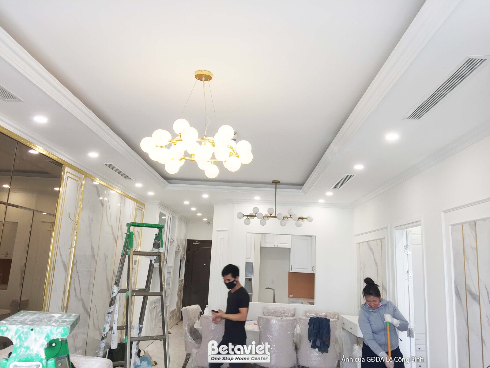 Góc nhìn tổng thể phòng khách từ cửa ra vào, cho thấy trần thạch cao giật cấp với đèn chùm lớn và hệ đèn âm trần. Tường đư…