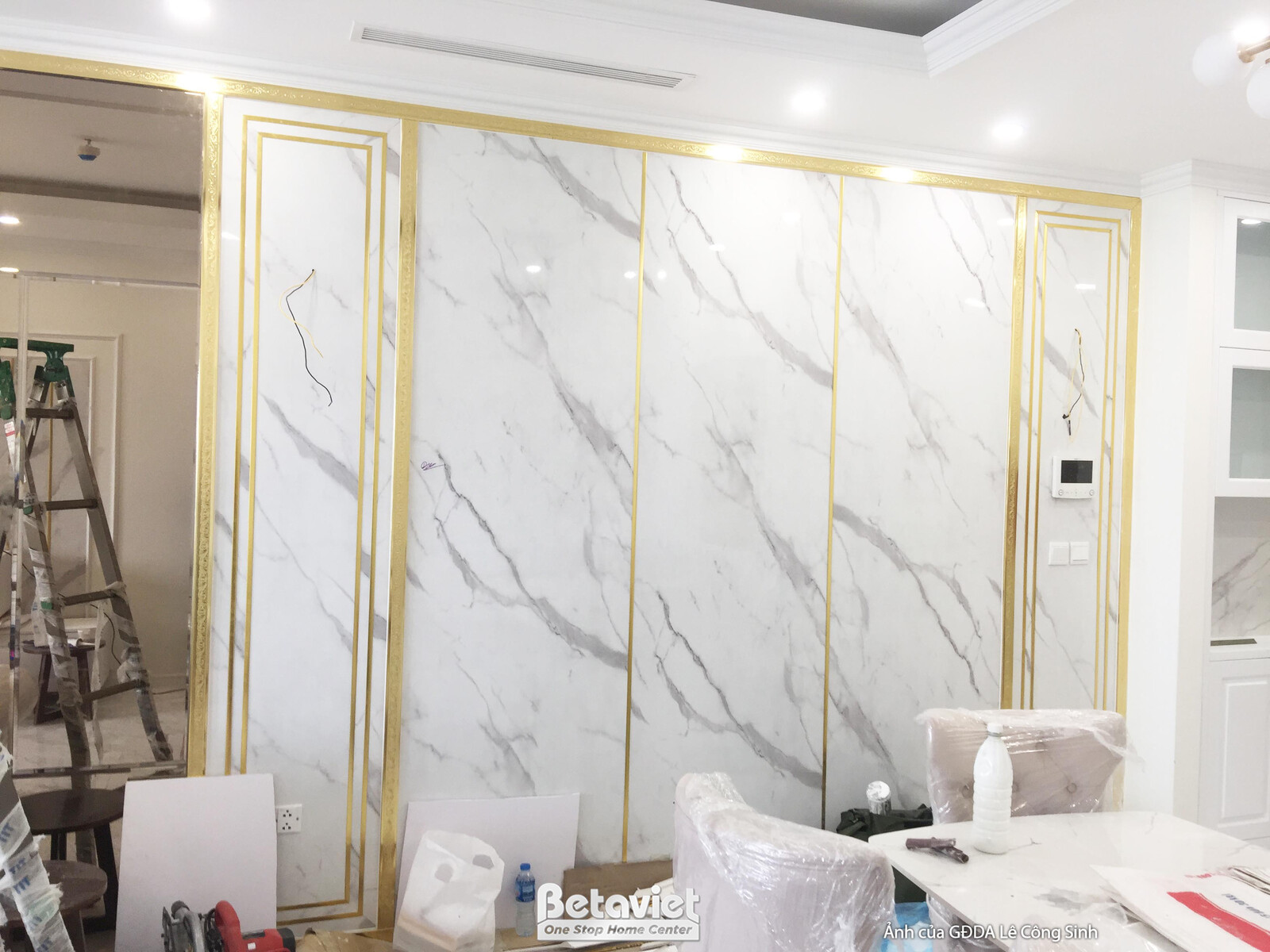 Không gian phòng khách với bức tường trang trí lớn ốp đá vân mây trắng và phào chỉ kết hợp nẹp vàng đồng, tạo điểm nhấn ấn…