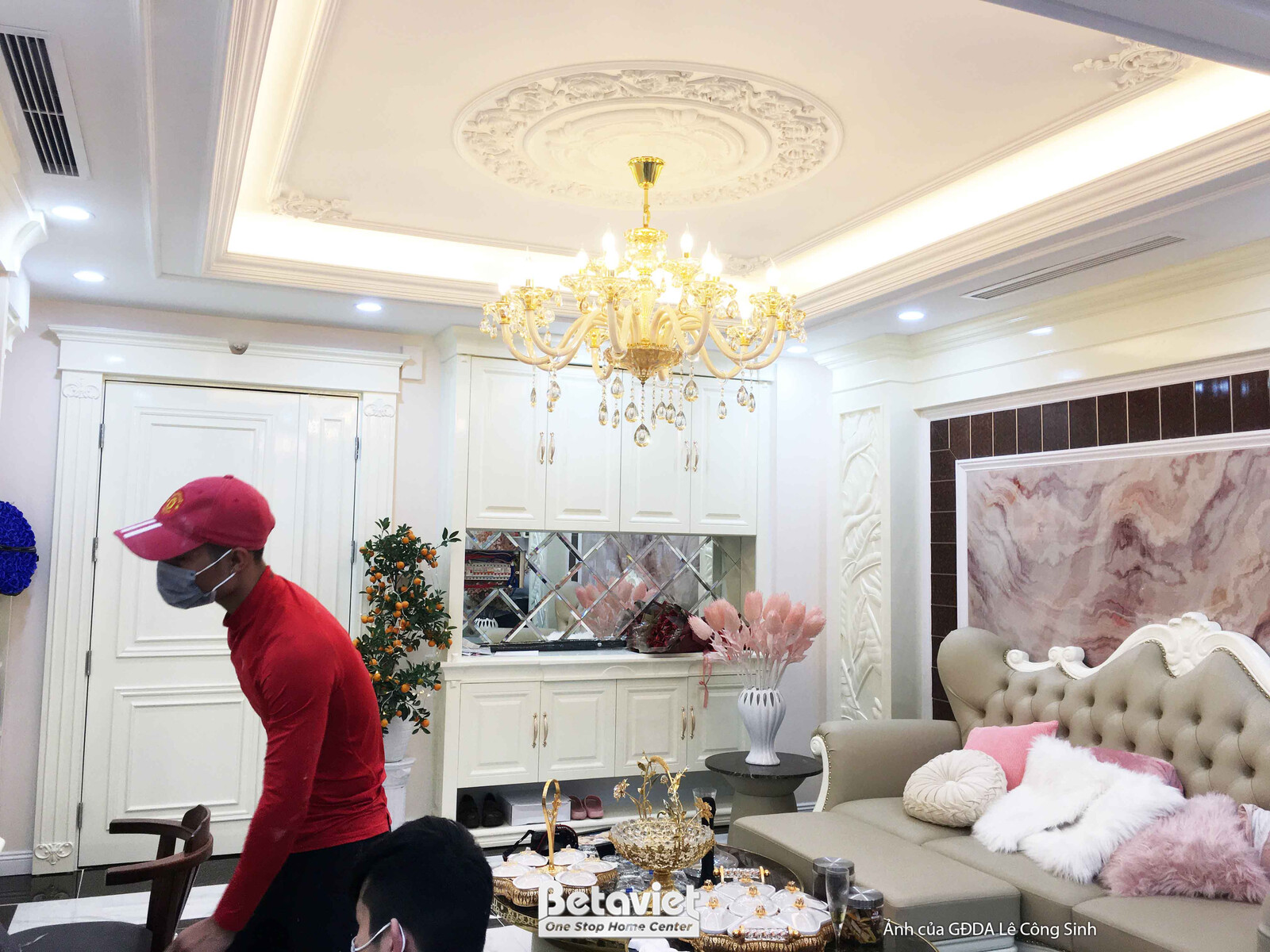 Toàn cảnh phòng khách tân cổ điển, với sofa chữ L màu be và bàn trà trung tâm. Phía đối diện là hệ tủ kệ màu trắng tinh tế…