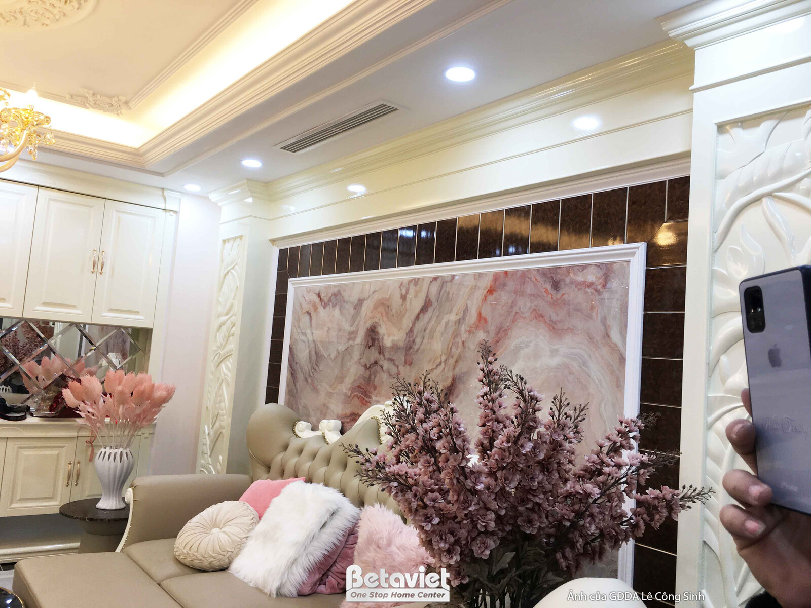 Cận cảnh khu vực sofa chữ L màu be với gối tựa hồng và trắng trong phòng khách. Điểm nhấn là bức tường ốp đá vân hồng nhạt…