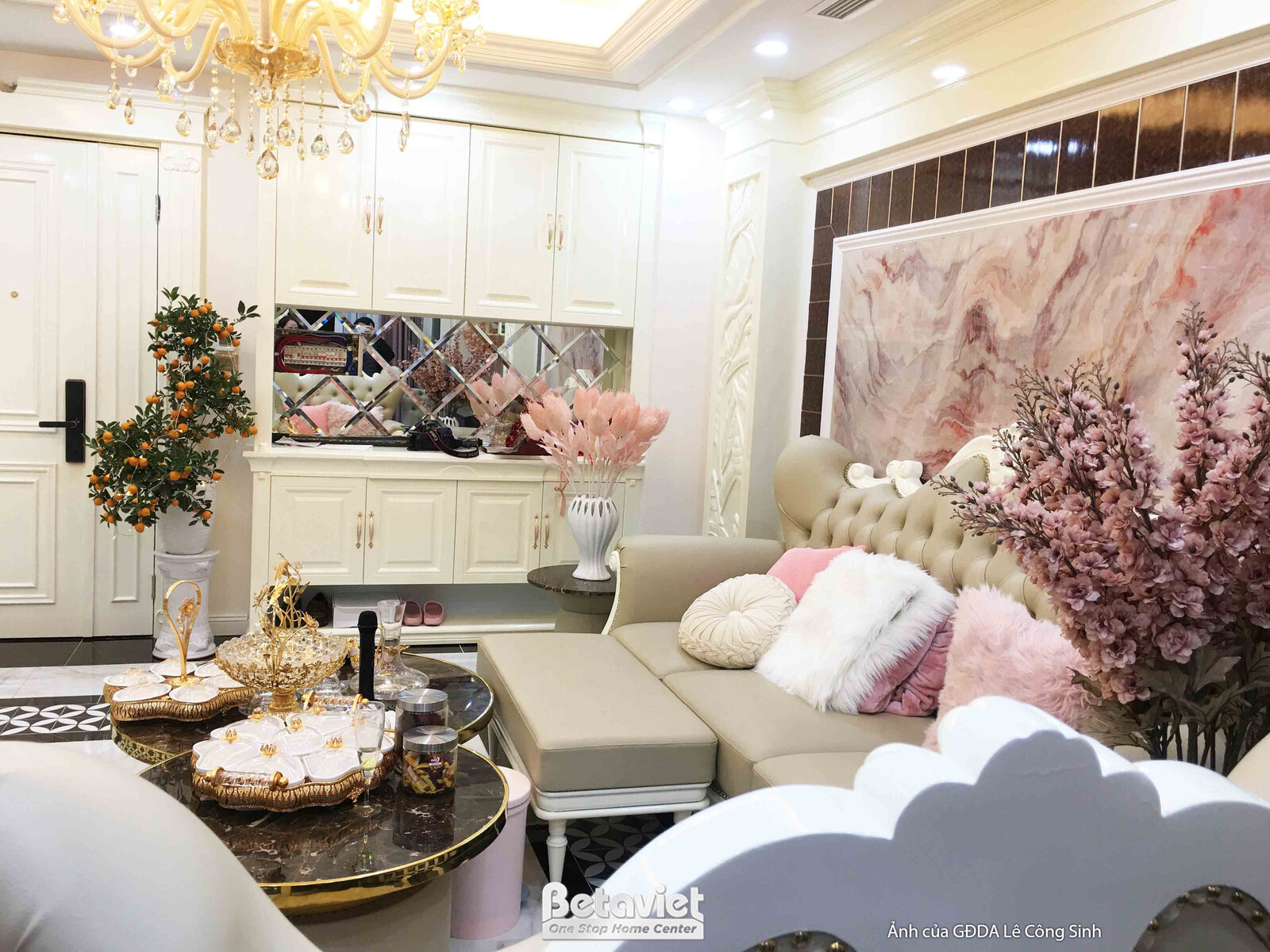 Góc phòng khách với sofa chữ L màu be, gối tựa mềm mại. Bàn trà mặt đá đen viền vàng đồng được trang trí tinh xảo. Tường ố…