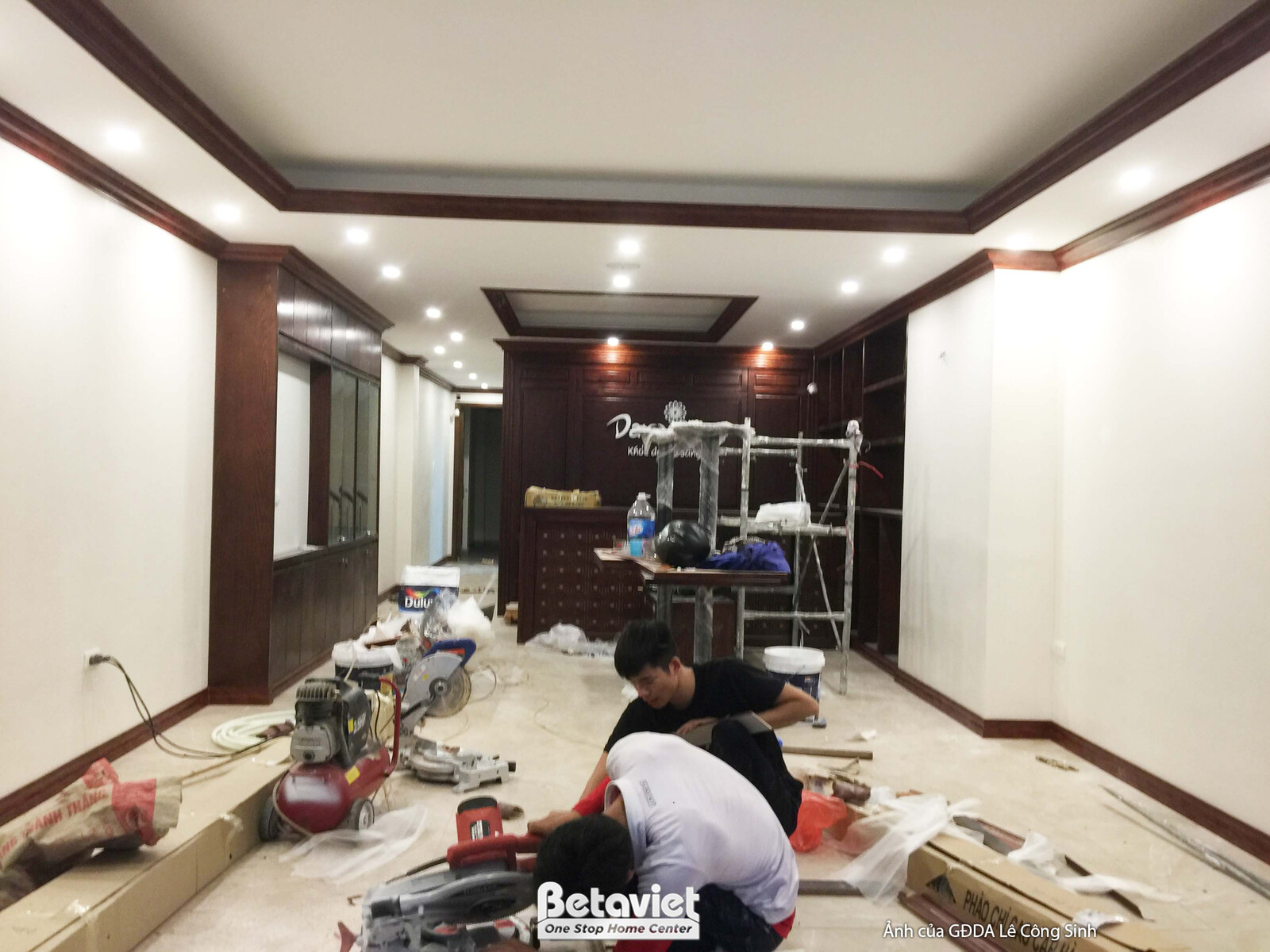 Góc nhìn tổng thể khu vực lễ tân và sảnh chờ spa, với các công nhân đang hoàn thiện sàn và lắp đặt. Bố cục rộng, cho thấy…