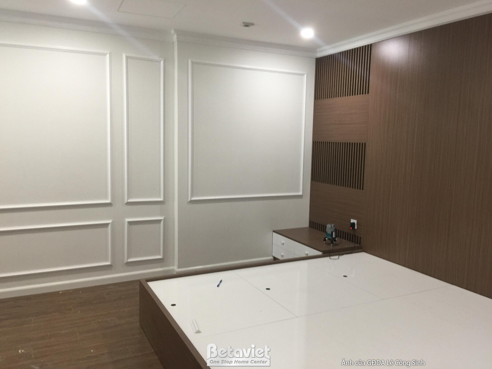 Phòng ngủ với mảng tường đầu giường ốp gỗ màu nâu ấm, kết hợp phào chỉ trắng ở các mảng tường còn lại. Giường ngủ màu trắn…