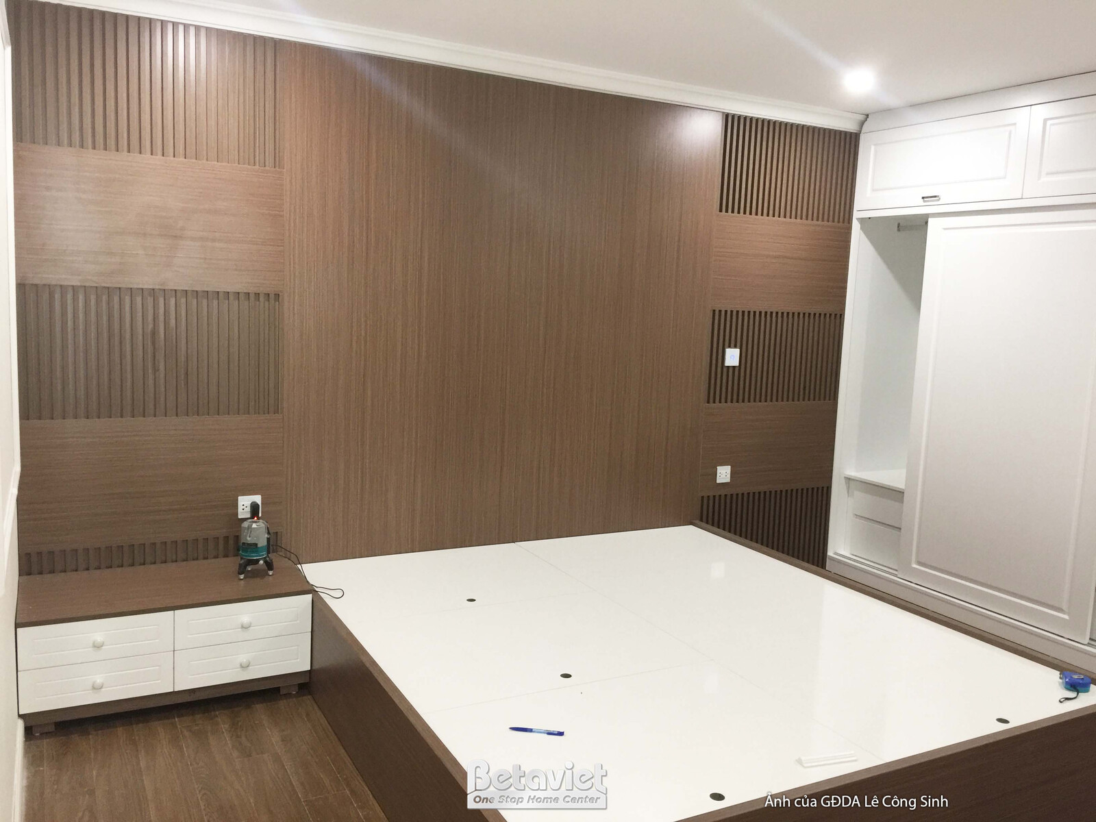 Toàn cảnh phòng ngủ với mảng tường đầu giường ốp gỗ màu nâu, giường ngủ màu trắng và tủ quần áo lớn màu trắng. Bàn đầu giư…