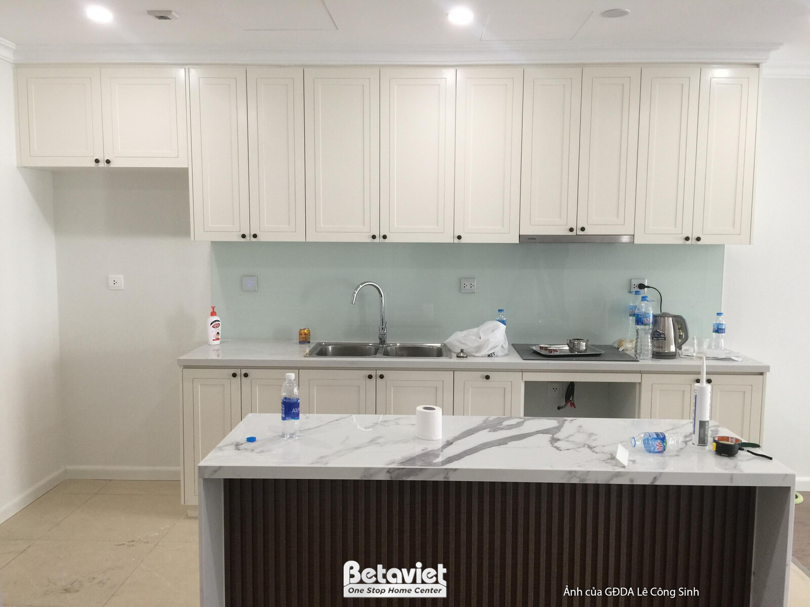 Khu vực bếp đã gần hoàn thiện với hệ tủ trên dưới màu trắng kem, mặt bàn đảo đá vân mây và lam gỗ trang trí. Kính ốp bếp m…