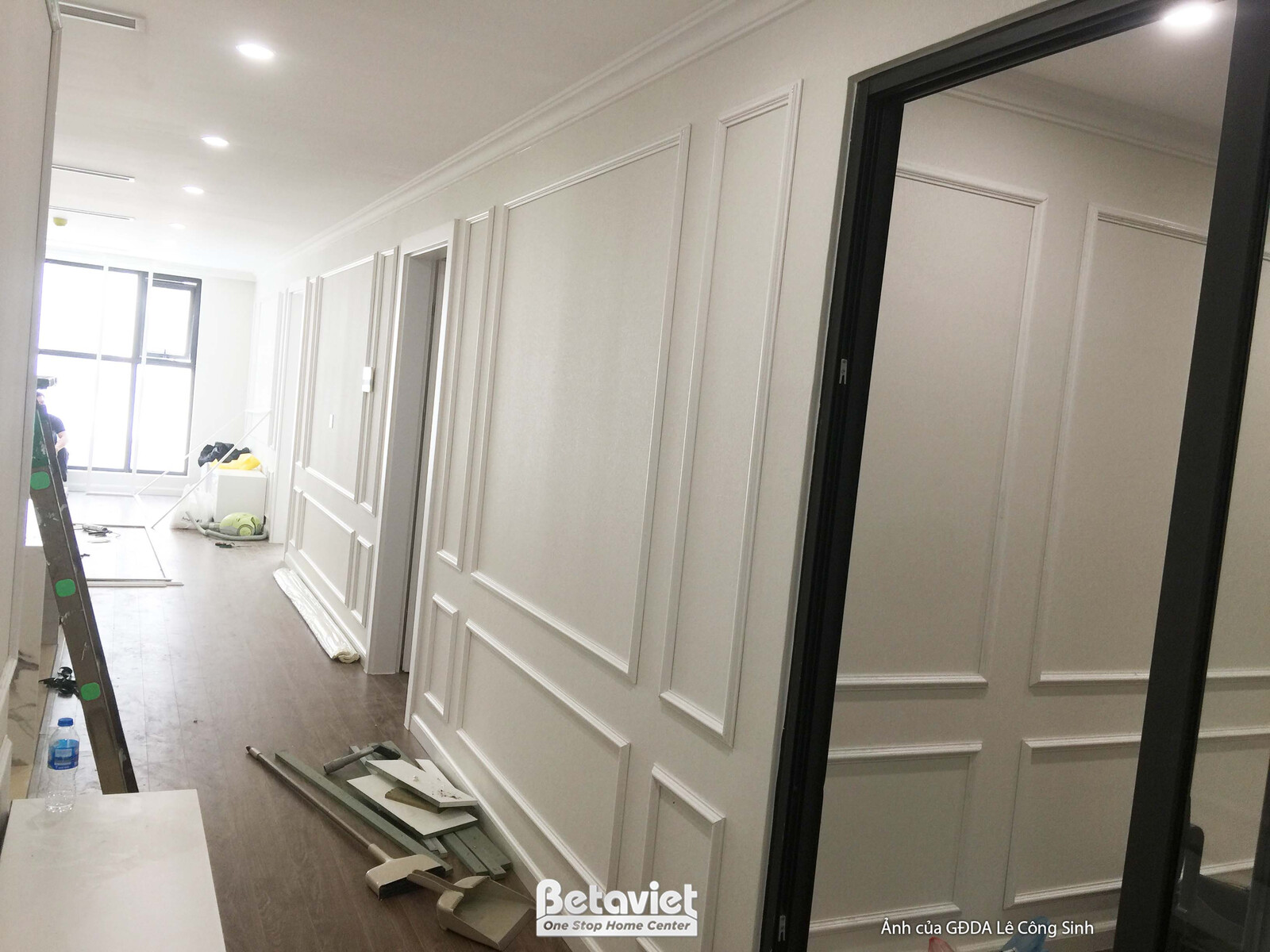 Hành lang dài với tường ốp phào chỉ trắng, cửa ra vào màu trắng. Một khung cửa lớn màu đen đang được lắp đặt, tạo điểm nhấ…