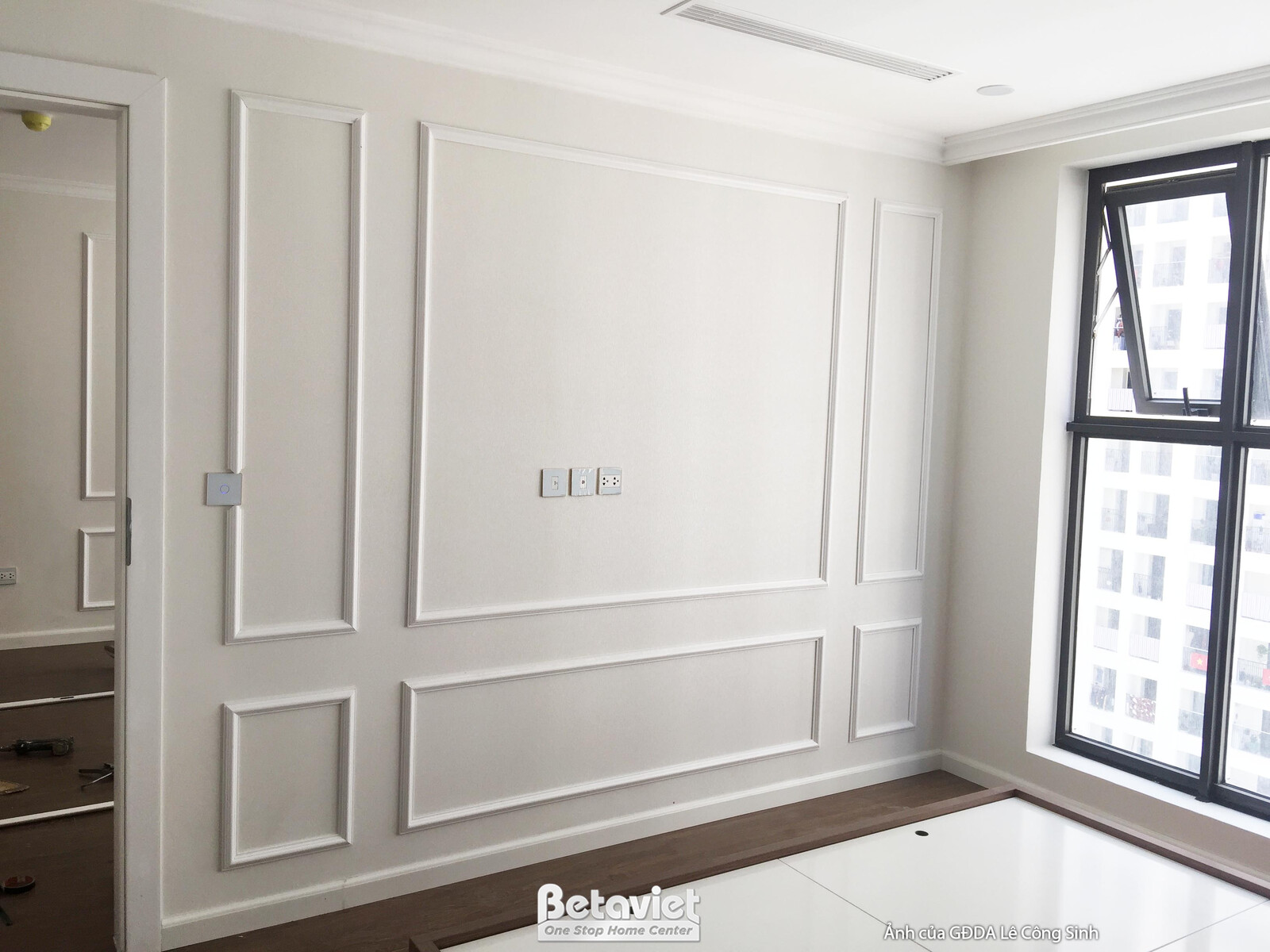 Góc phòng với tường ốp phào chỉ trắng, cửa sổ lớn đón ánh sáng tự nhiên. Sàn nhà lát gỗ màu nâu ấm. Không gian đơn giản nh…
