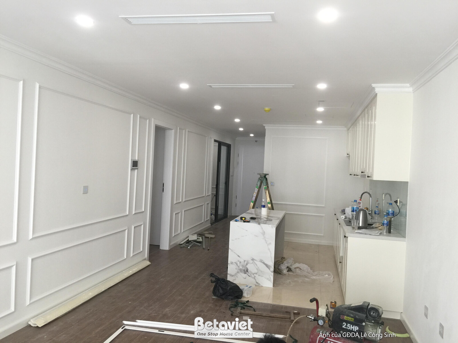 Toàn cảnh không gian phòng khách và bếp nhìn từ phía cửa sổ, đang trong giai đoạn hoàn thiện. Tường ốp phào chỉ trắng, khu…