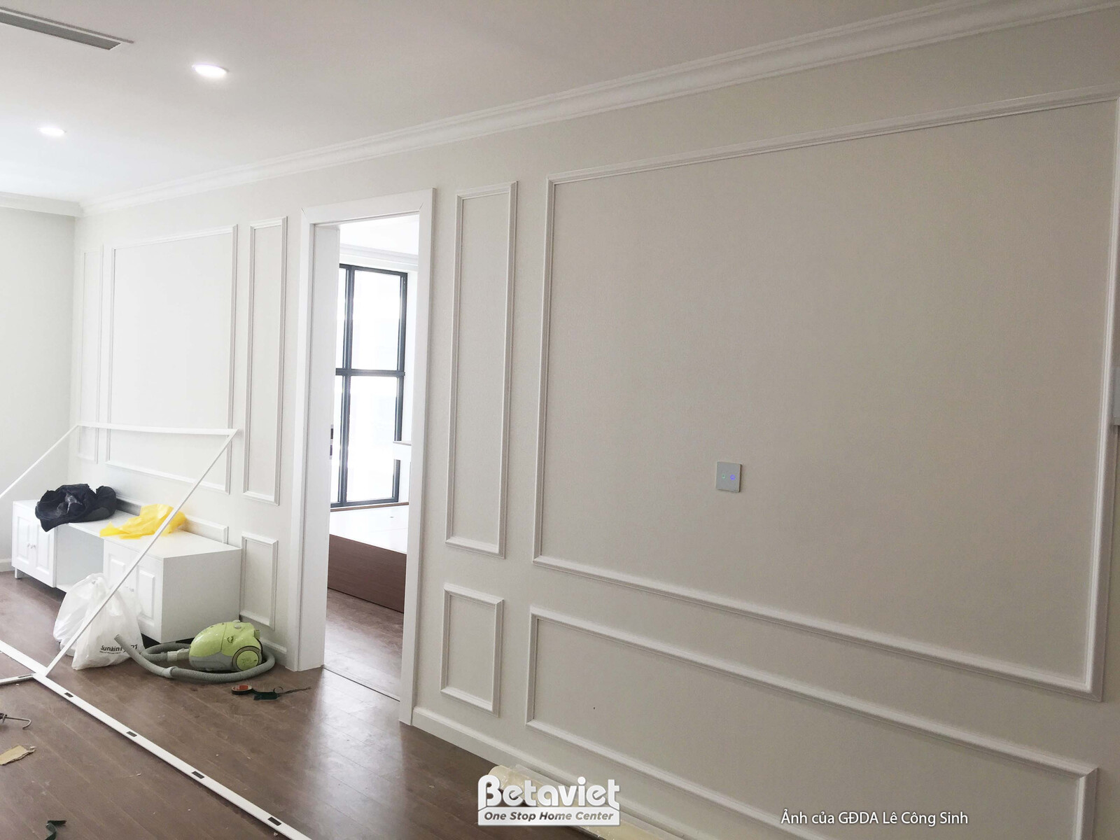 Góc phòng khách hoặc khu vực sinh hoạt chung, tường ốp phào chỉ trắng, cửa ra vào màu trắng. Sàn nhà đang được hoàn thiện…
