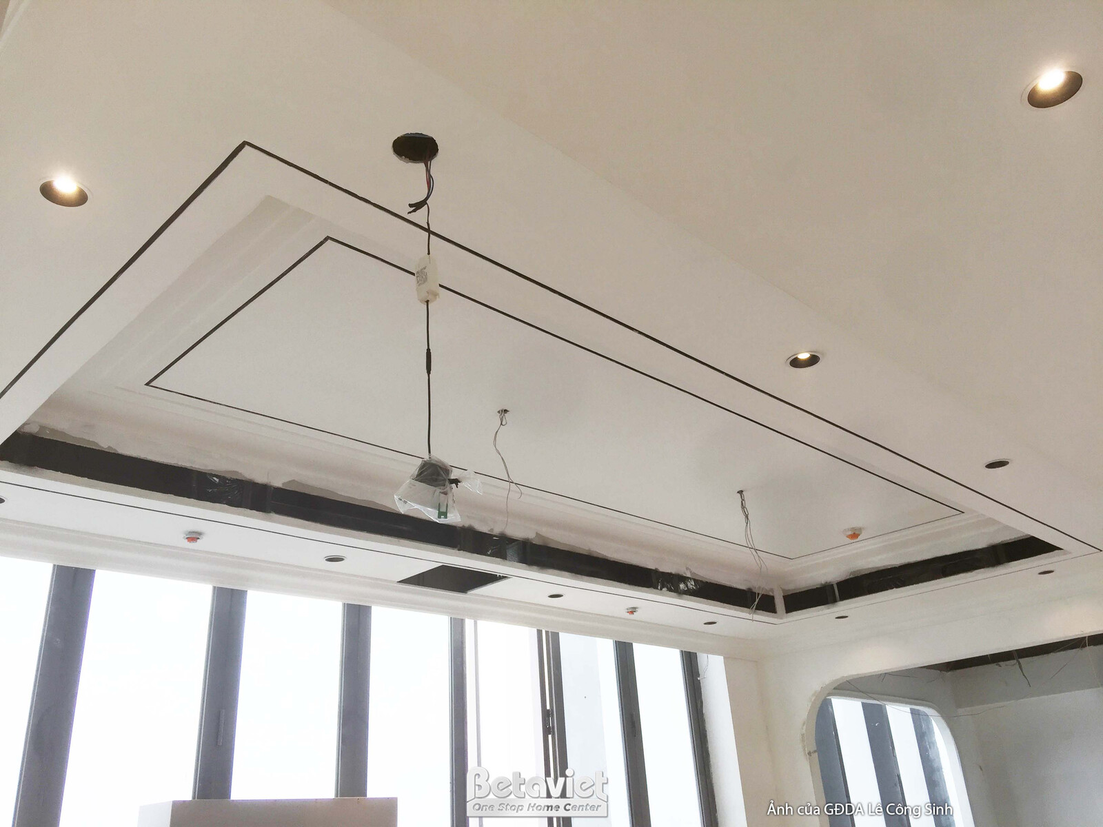 Trần thạch cao giật cấp với phào chỉ và hệ thống đèn âm trần đang được lắp đặt. Cửa sổ lớn cung cấp ánh sáng tự nhiên, tạo…